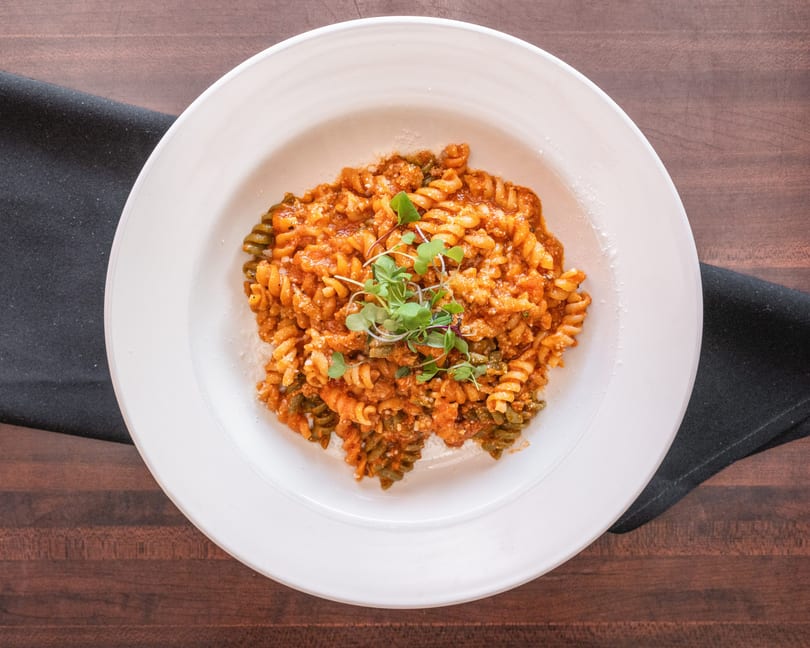 A white bowl of fusilli pasta with red sauce, cheese, and microgreens on a wooden table.