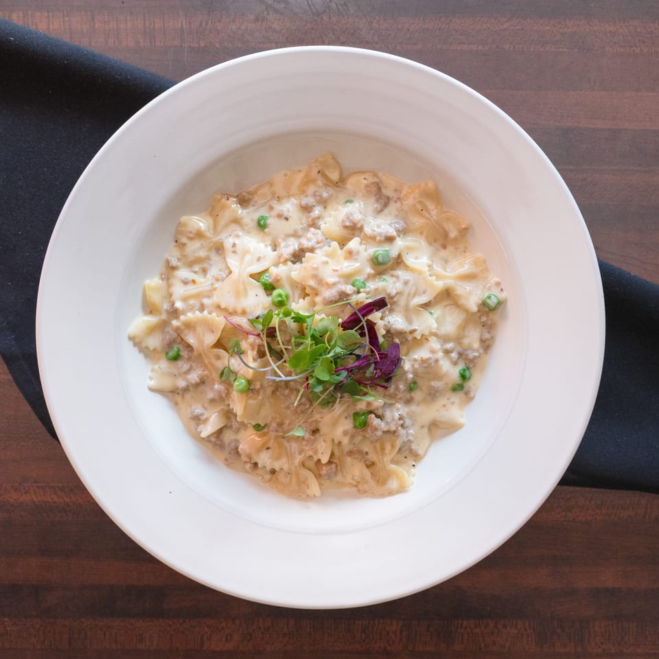 Creamy farfalle pasta with ground meat, peas, and microgreens in a white bowl on a wooden table.
