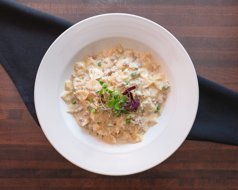 Creamy farfalle pasta with ground meat, peas, and microgreens in a white bowl on a wooden table.