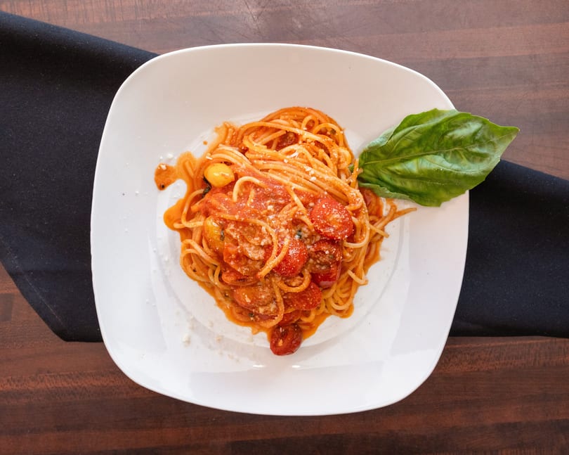 Spaghetti with tomato sauce, cherry tomatoes, Parmesan cheese, and a basil leaf on a white plate.