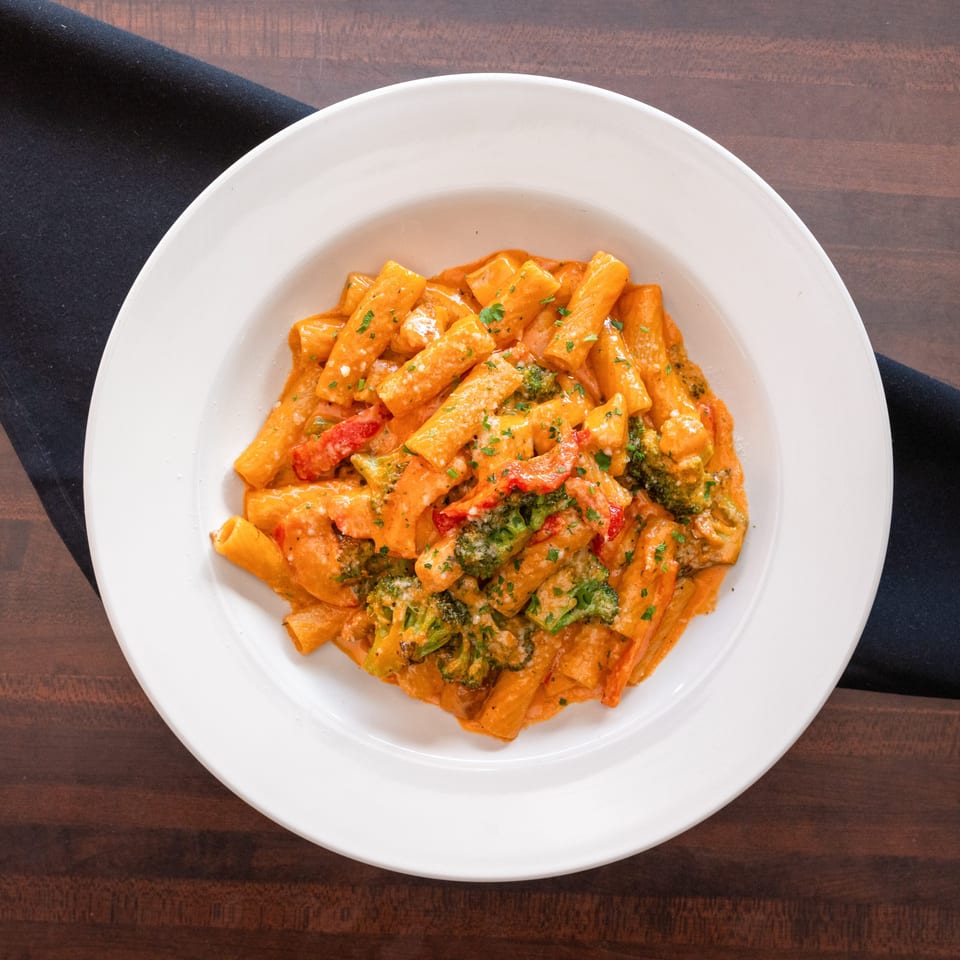 Rigatoni pasta with creamy tomato sauce, broccoli, red peppers, and parsley in a white bowl.