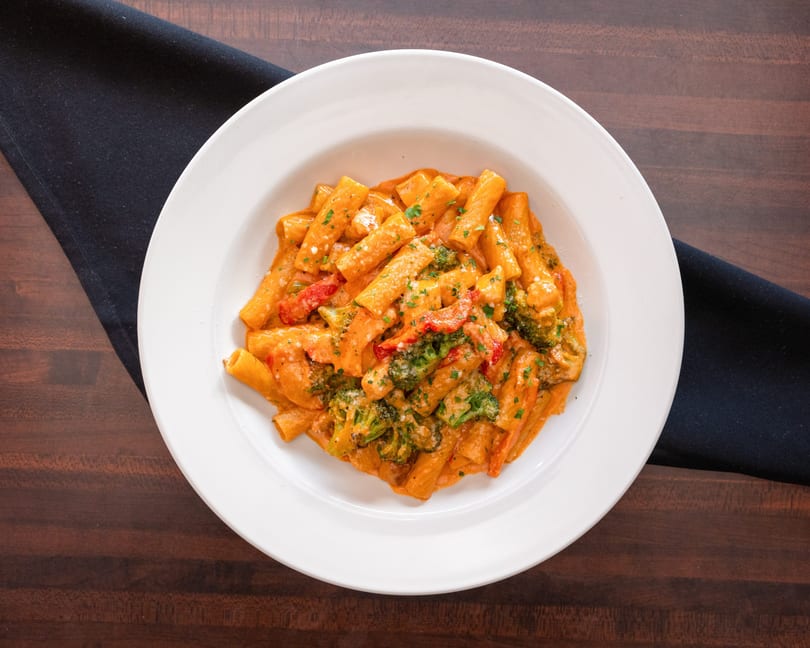 Rigatoni pasta with creamy tomato sauce, broccoli, red peppers, and parsley in a white bowl.
