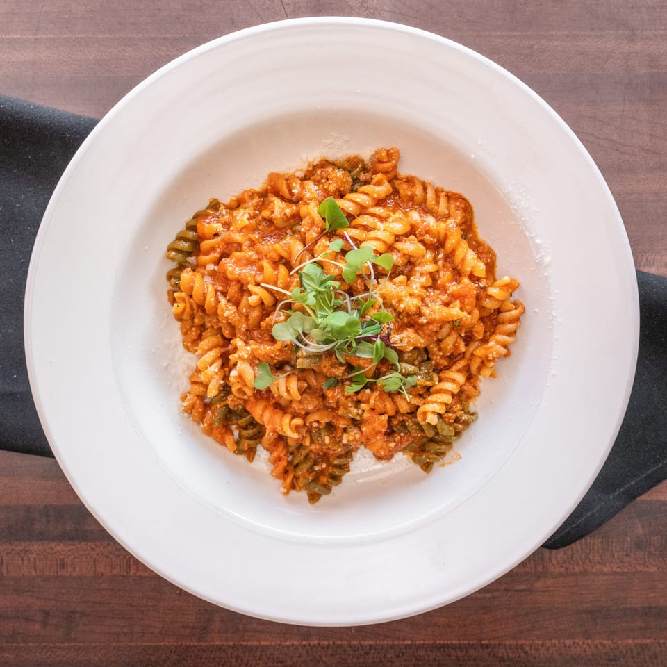 A white bowl of fusilli pasta with red sauce, cheese, and microgreens on a dark wooden table.