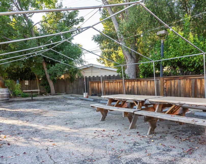 Outdoor dining area with picnic tables under a metal frame with string lights, surrounded by trees.
