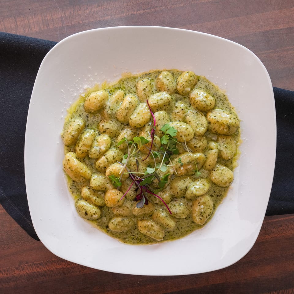 A white square plate filled with gnocchi in green pesto sauce, garnished with microgreens.