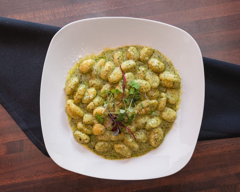 A white square plate filled with gnocchi in green pesto sauce, garnished with microgreens.