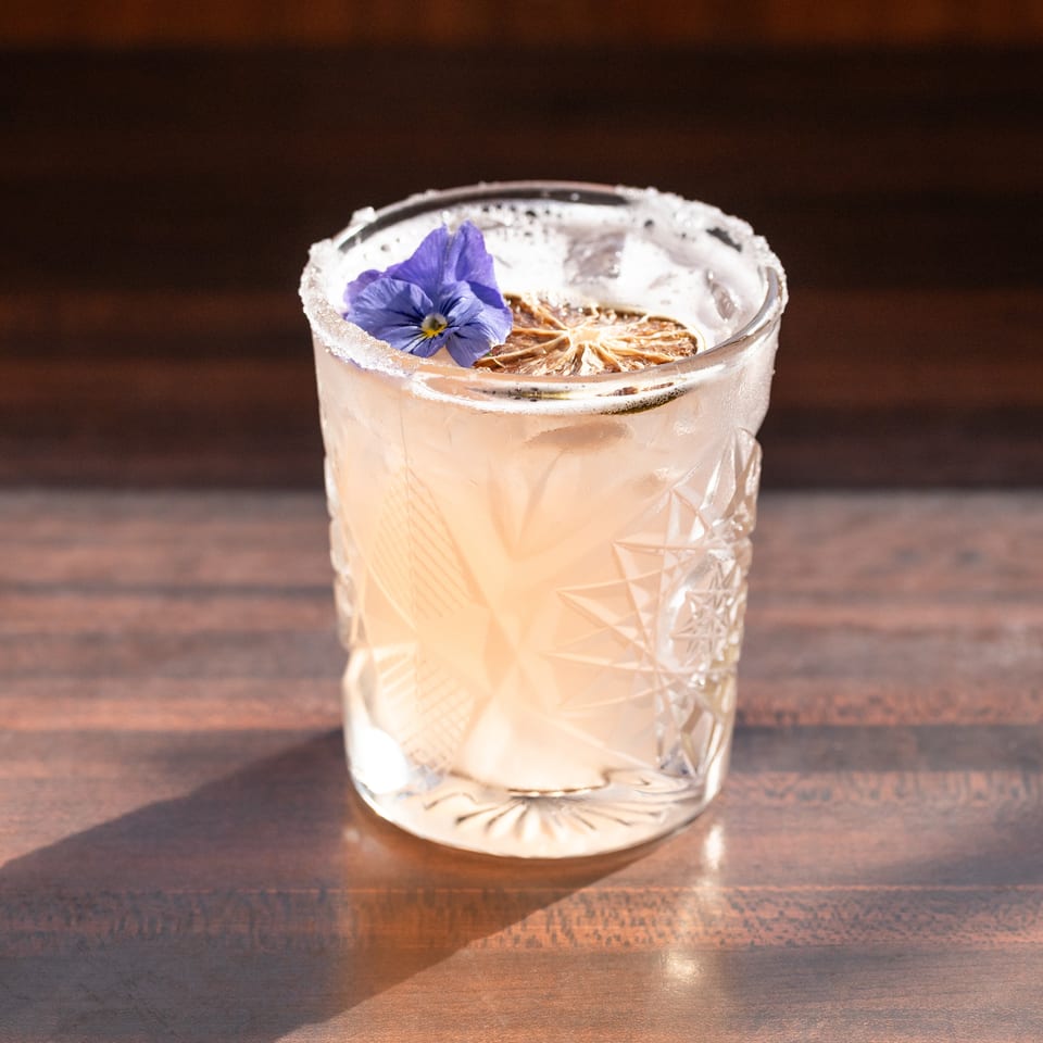 A light pink cocktail in a textured glass, garnished with a dried lime and purple flower.