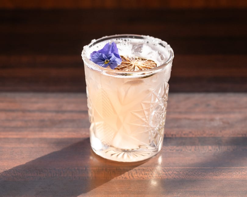 A light pink cocktail in a textured glass, garnished with a dried lime and purple flower.