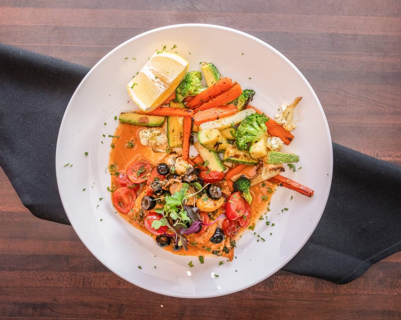 Grilled salmon with roasted vegetables, cherry tomatoes, olives, and a lemon wedge.