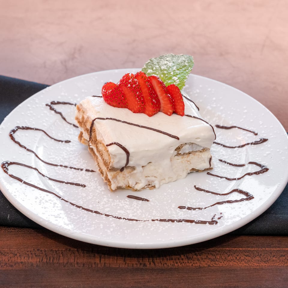 Tiramisu dessert with whipped cream, sliced strawberries, chocolate drizzle, and mint on a white plate.