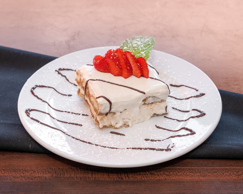 Tiramisu dessert with whipped cream, sliced strawberries, chocolate drizzle, and mint on a white plate.