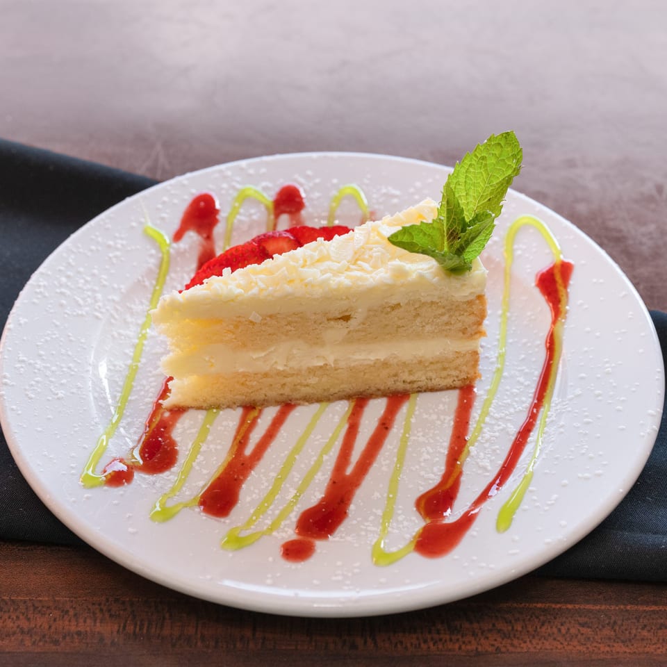 Slice of white cake with frosting, strawberries, mint, and colorful drizzles on a white plate.