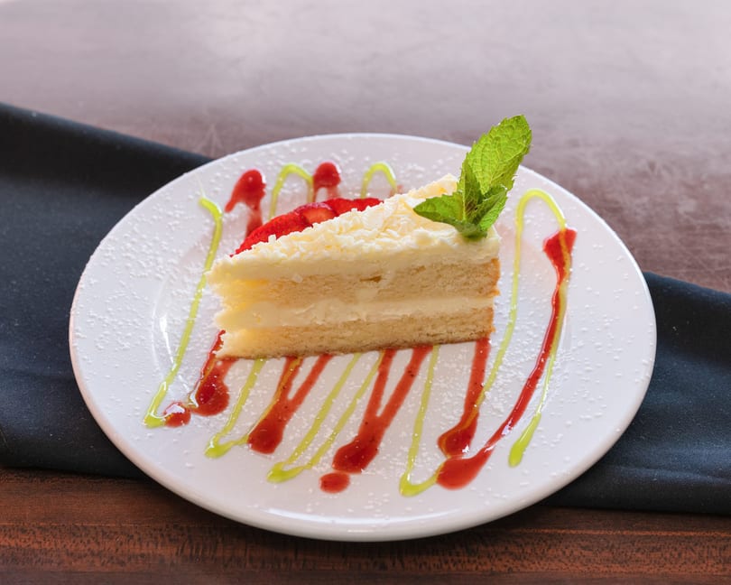 Slice of white cake with frosting, strawberries, mint, and colorful drizzles on a white plate.