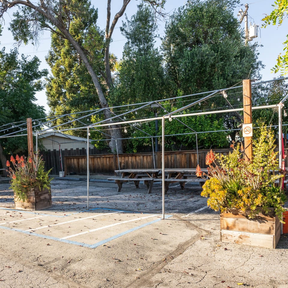 Outdoor dining area with picnic tables under a metal frame with string lights, surrounded by trees.