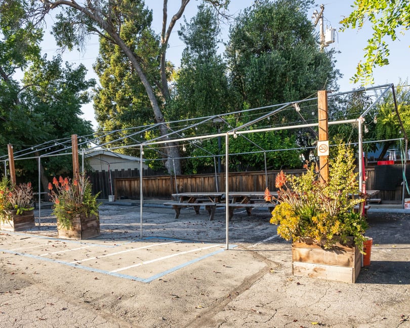 Outdoor dining area with picnic tables under a metal frame with string lights, surrounded by trees.